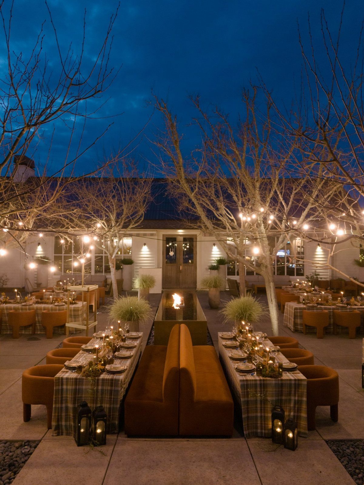 Long dining tables with plaid tablecloths and lanterns under lit bare trees.