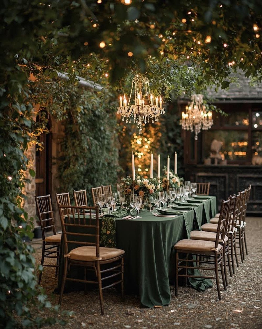 Forest green dinner table with gold chairs and chandeliers under leafy trees