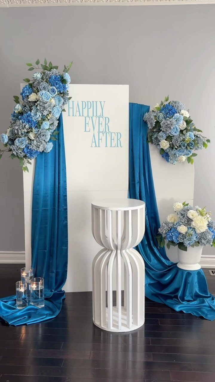Blue and white wedding backdrop with satin drapes, floral arrangements, and a white pedestal.