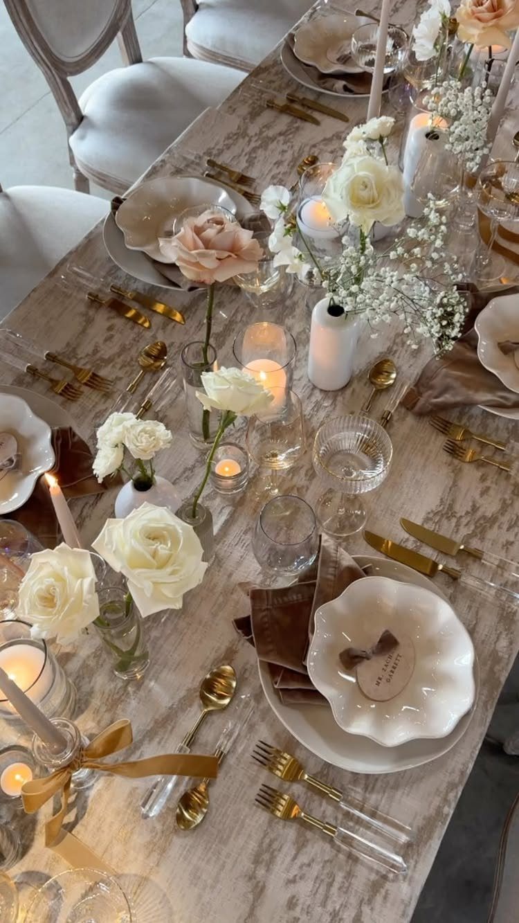 Wedding table setting with gold cutlery, white roses, and glowing candles on marble.