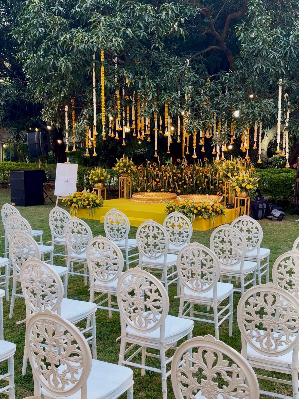 Outdoor wedding ceremony setup with white chairs, yellow floral decor, and hanging garlands.